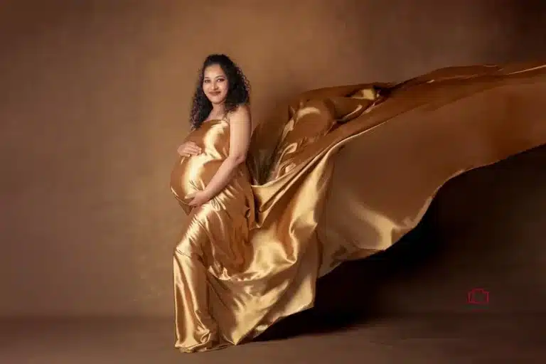 A radiant mother-to-be in a golden satin gown with a flowing train, beautifully captured in a maternity studio photoshoot