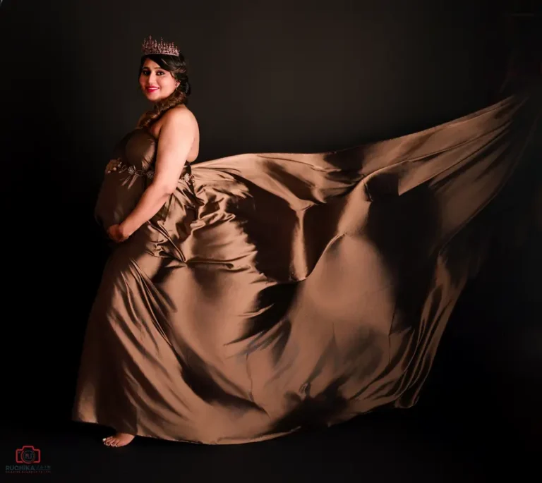 A glowing mother-to-be in a champagne satin gown with a flowing train and tiara poses gracefully in a maternity studio photoshoot. This maternity photography session captures regal beauty and confidence