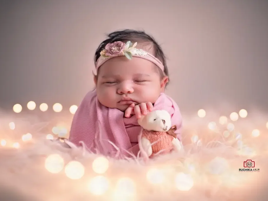 Newborn baby in froggy pose wrapped in a pink blanket with a floral headband, peacefully sleeping while holding a small teddy bear, surrounded by soft bokeh lights