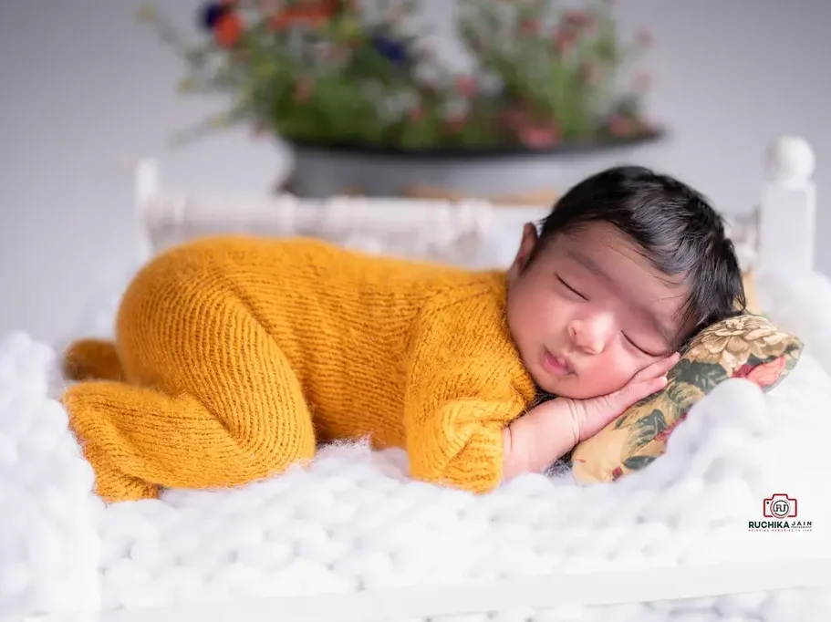 Newborn baby in a mustard yellow outfit, peacefully sleeping on a small white prop bed with a floral pillow and blurred floral background