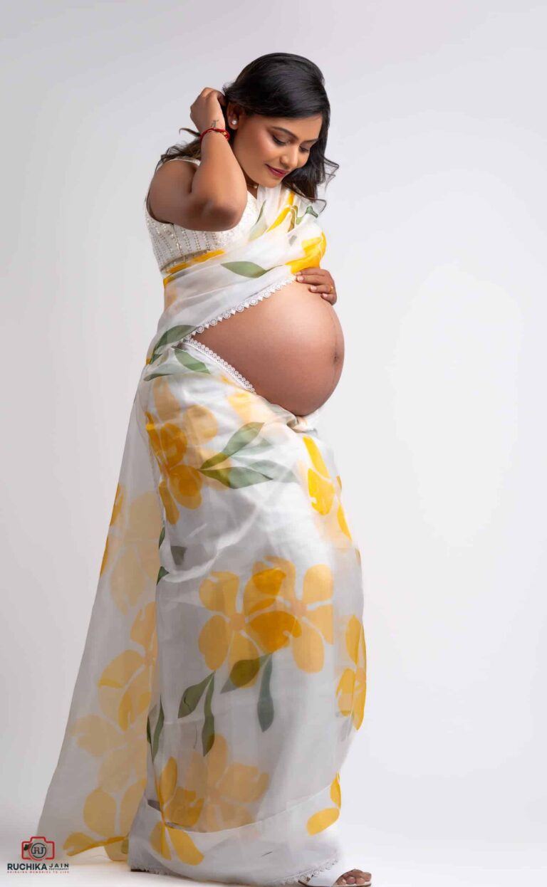 A glowing mother-to-be in a floral saree gently cradles her baby bump during a maternity studio photoshoot. This elegant pregnancy photography session captures joy, beauty, and anticipation