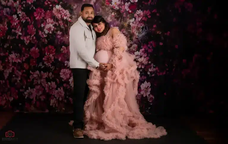 A loving expecting couple poses in a dreamy maternity studio photoshoot with a floral backdrop. The mother-to-be stuns in a soft pink ruffled gown, embracing the joy of pregnancy