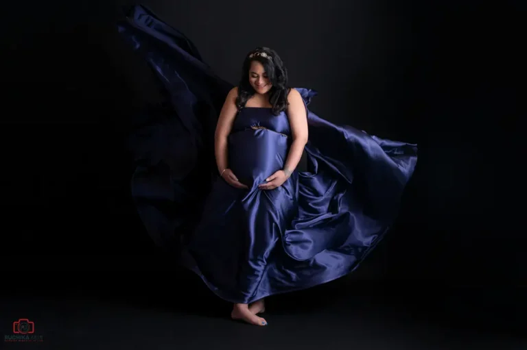 A glowing mother-to-be in a dramatic navy satin gown cradles her baby bump during a maternity studio photoshoot. The flowing fabric adds elegance, capturing the beauty and grace of pregnancy
