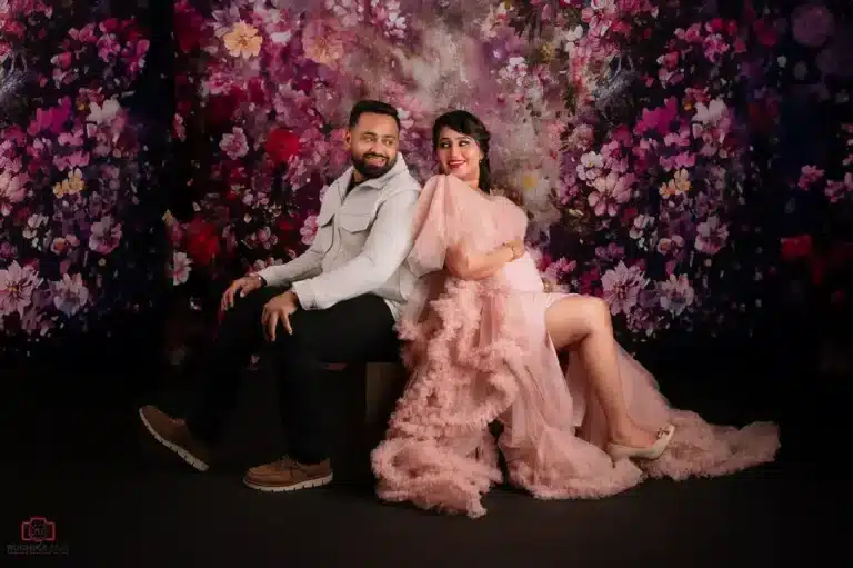 A joyful expecting couple poses back-to-back in a maternity studio photoshoot with a vibrant floral backdrop. The mother-to-be looks radiant in a flowing pink ruffled gown, embracing the excitement of pregnancy
