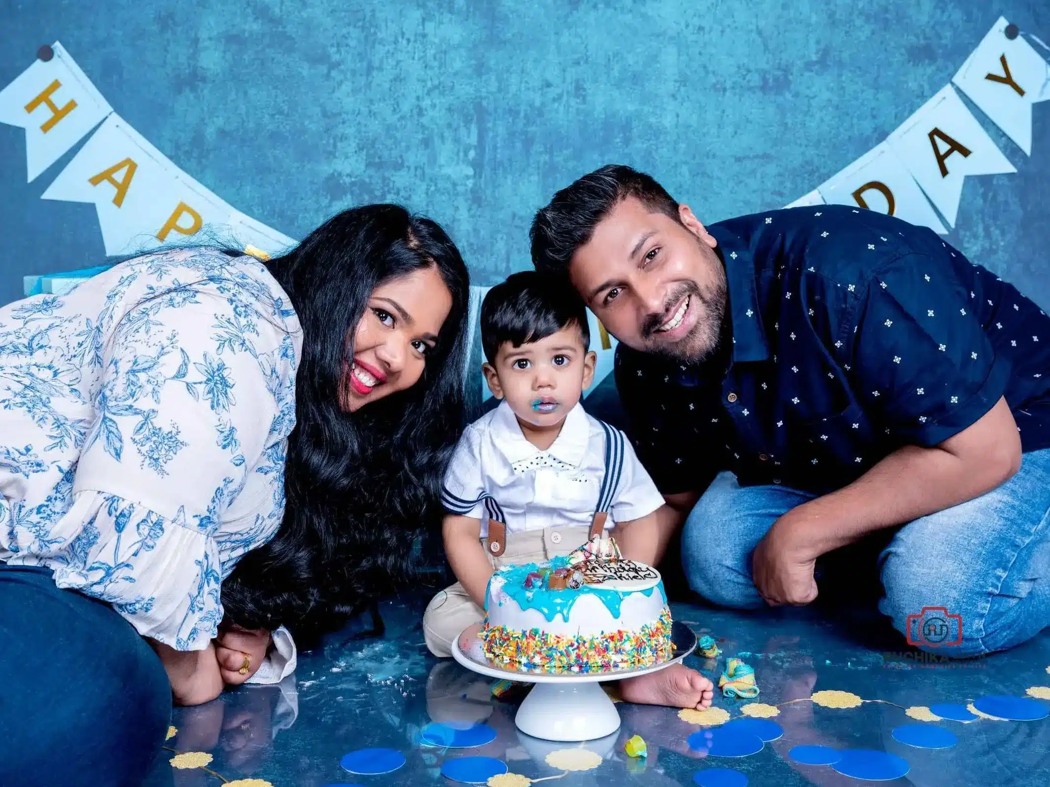 Joyful Cake Smash Celebration: Parents join in the fun as their child gleefully smashes a cake at the center of a blue-themed studio during a cake smash and splash photography session